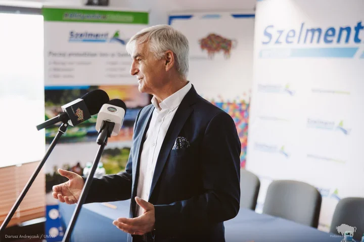 Mężczyzna w ciemnej marynarce przemawia do mikrofonów na tle ścianek z napisem Szelment.