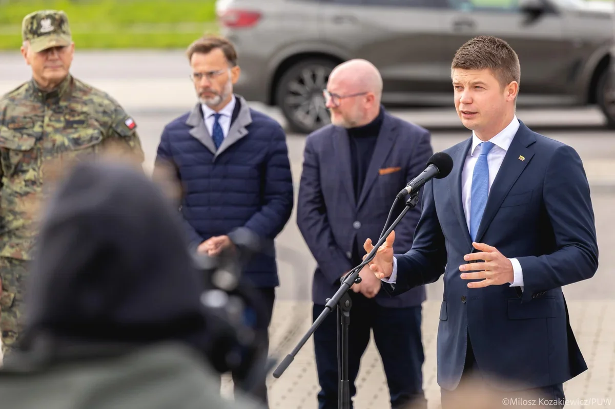 Mężczyzna w garniturze przemawia do mikrofonu na zewnątrz, za nim stoją trzej mężczyźni, w tym żołnierz w mundurze.