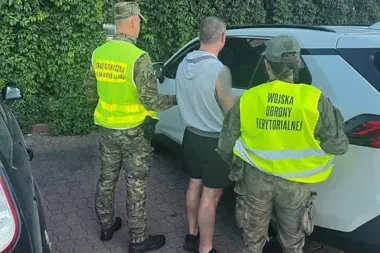 Funkcjonariusz Straży Granicznej i żołnierz Wojsk Obrony Terytorialnej zatrzymują mężczyznę przy białym samochodzie na brukowanym podjeździe.