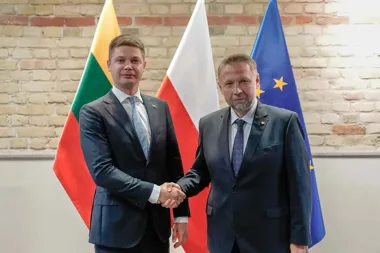 Dwaj mężczyźni w garniturach witają się uściskiem dłoni przed flagami Litwy, Polski i Unii Europejskiej.