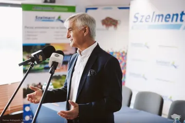 Mężczyzna w ciemnej marynarce przemawia do mikrofonów na tle ścianek z napisem Szelment.