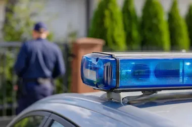 Policyjny radiowóz z niebieskim światłem ostrzegawczym na dachu, w tle rozmazany funkcjonariusz w mundurze.