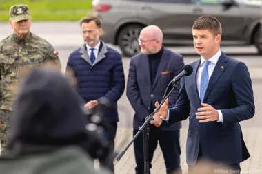 Mężczyzna w garniturze przemawia do mikrofonu na zewnątrz, za nim stoją trzej mężczyźni, w tym żołnierz w mundurze.